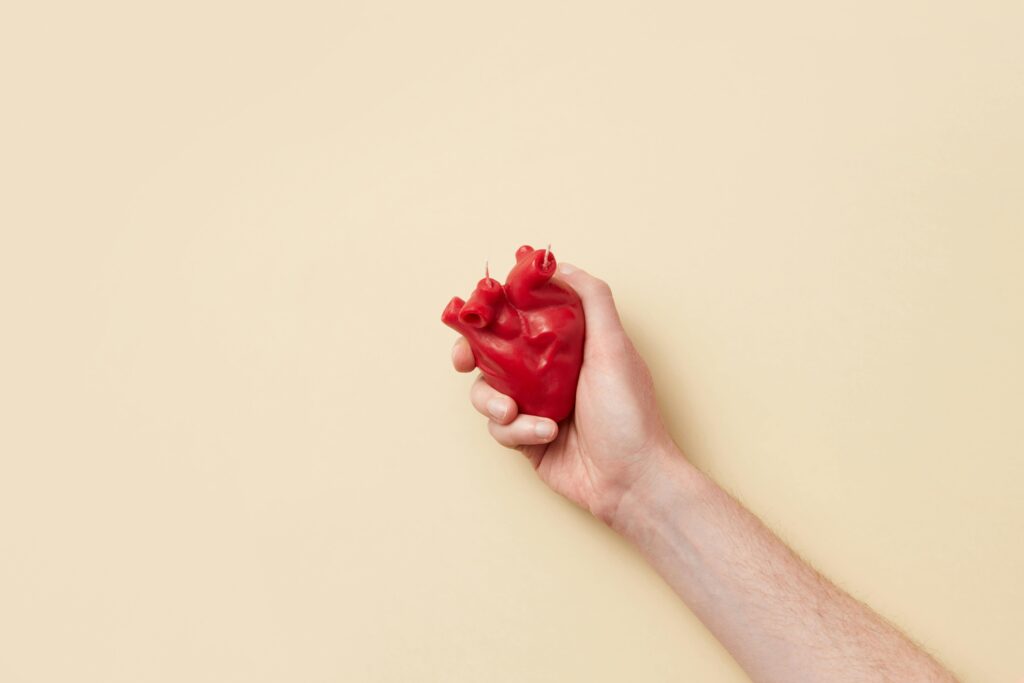A realistic red anatomical heart model held firmly in a hand against a beige background.